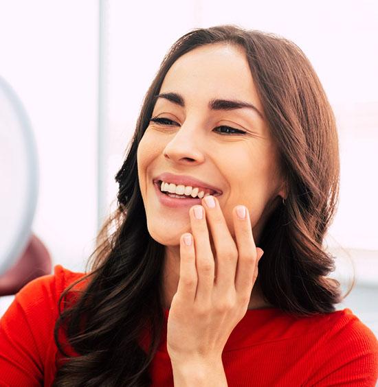 A young woman wearing a red blouse admires her smile in the mirror after receiving dental cleanings in Rockledge